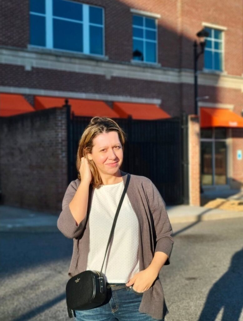 Woman wearing a white shirt, grey cardigan, and jeans standing outdoors.