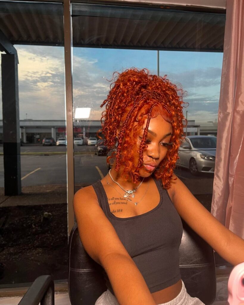 Woman with bright orange-red braided bob hairstyle wearing a grey crop top.