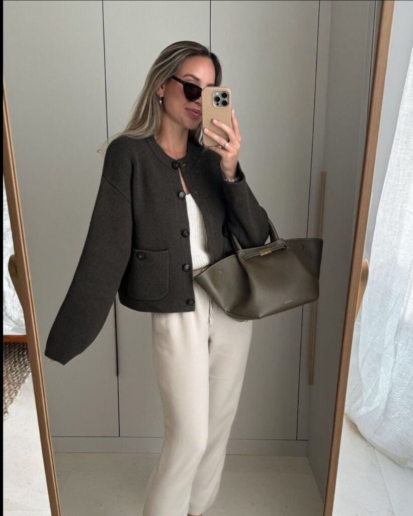 Woman in olive green cardigan and cream trousers holding a matching leather handbag.
