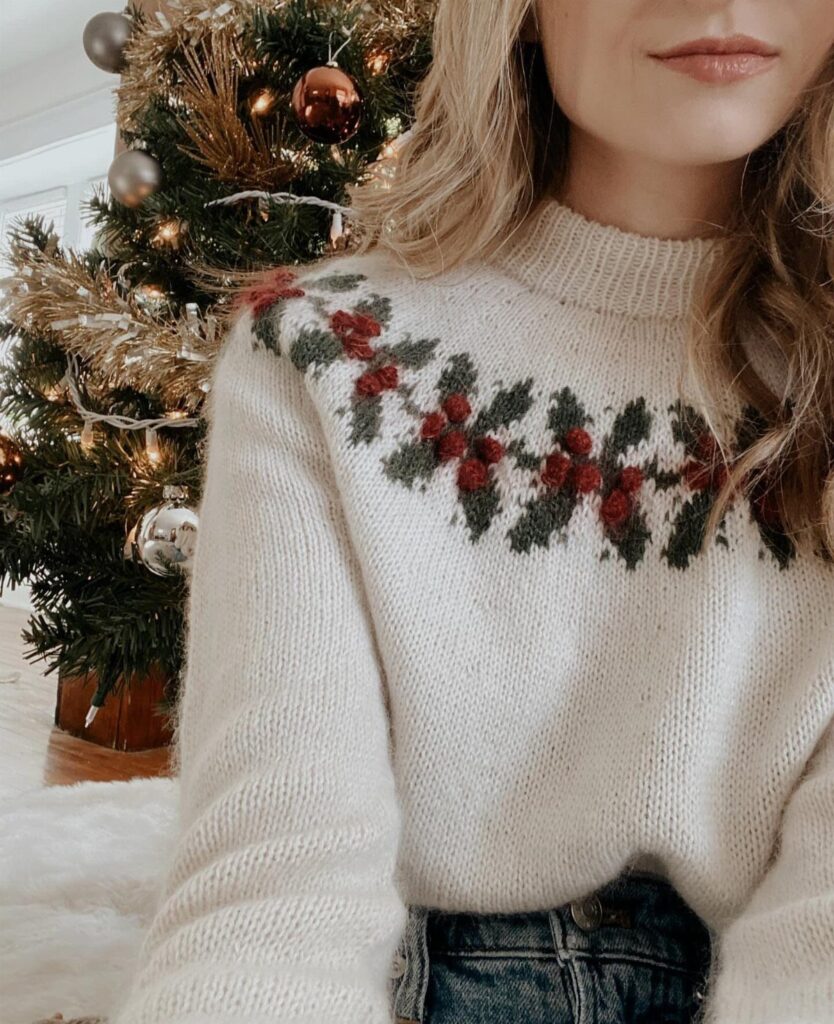 Woman wearing a cream knit sweater with holly berries next to a Christmas tree