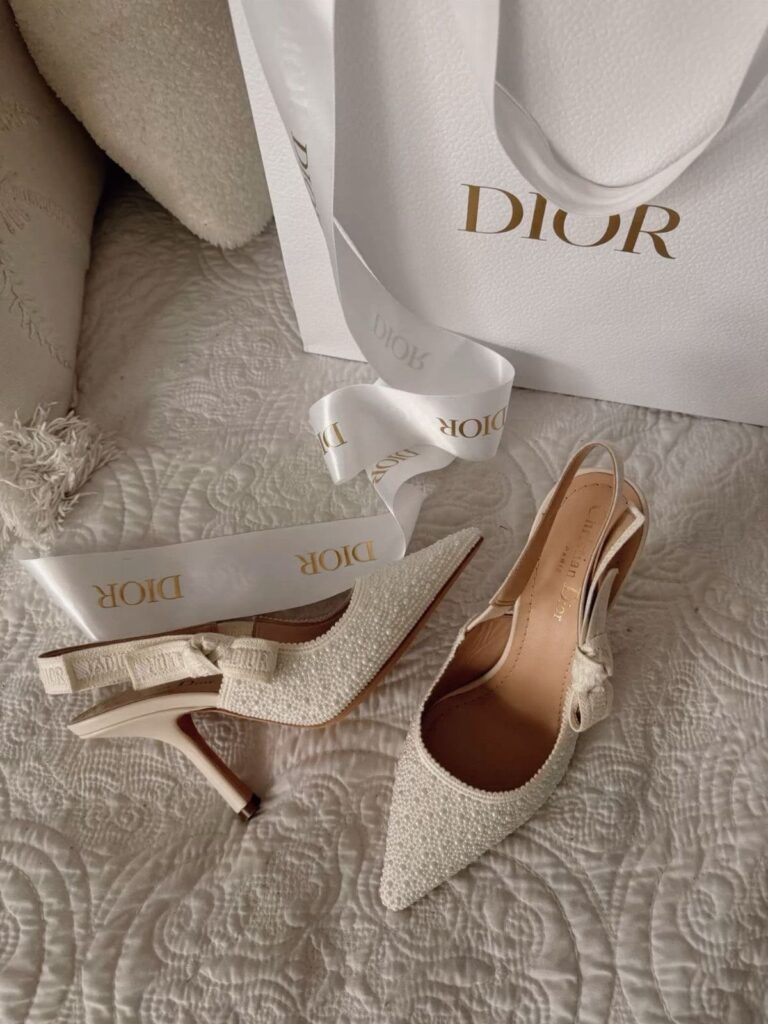 White sparkling Dior pointed-toe heels with bows next to a Dior shopping bag.