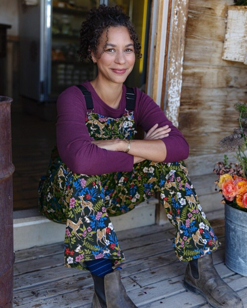 Woman wearing botanical print overalls and a purple shirt sitting on a porch.