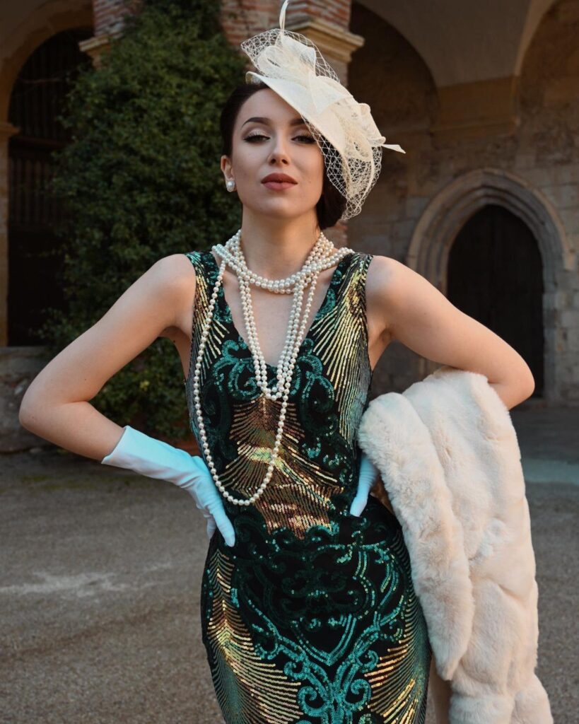 Woman in a green sequined gown with pearls and a white fascinator hat.