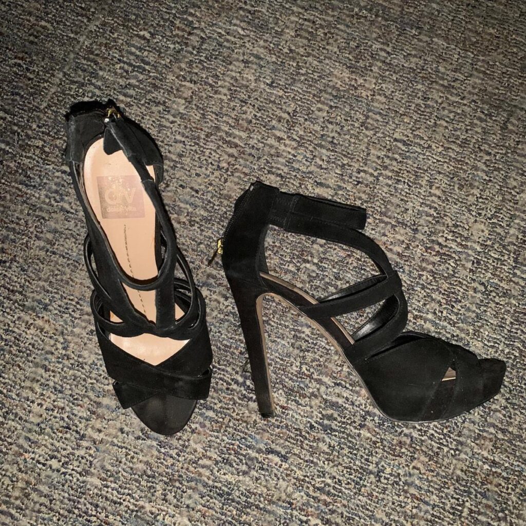 Black suede strappy high heels with ankle straps on a grey carpet.