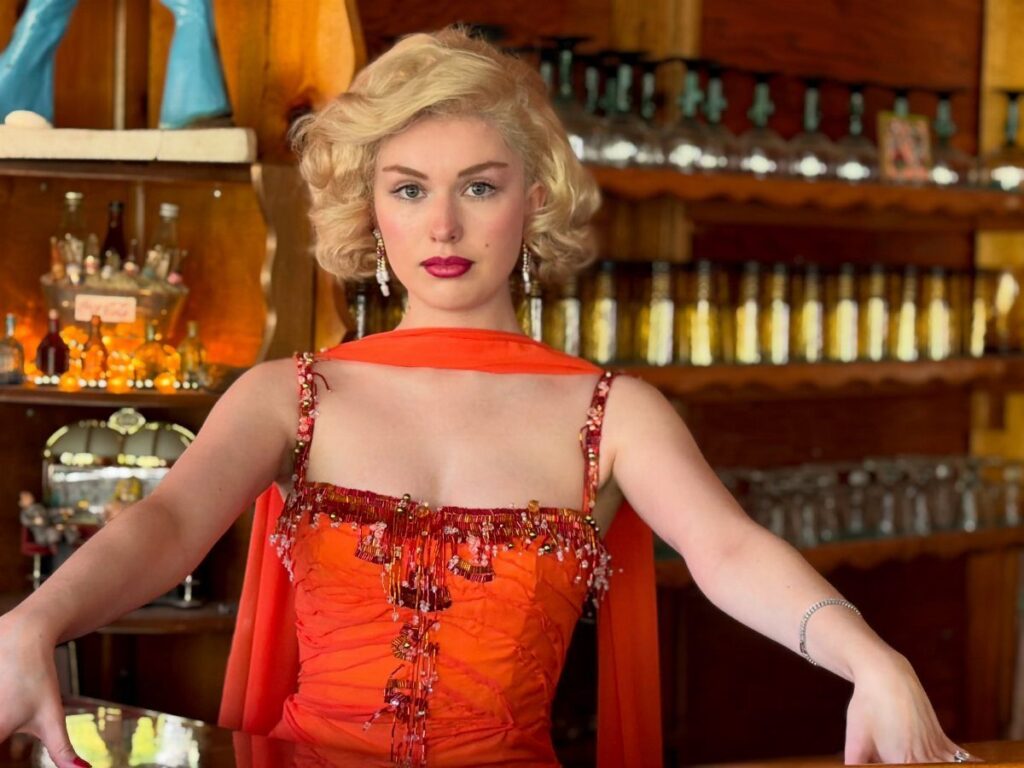 Blonde woman in a vintage orange beaded gown posing in a rustic bar.