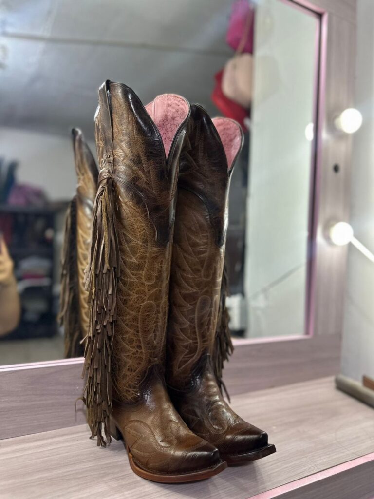 Brown leather cowboy boots with fringe standing on a wooden vanity table.